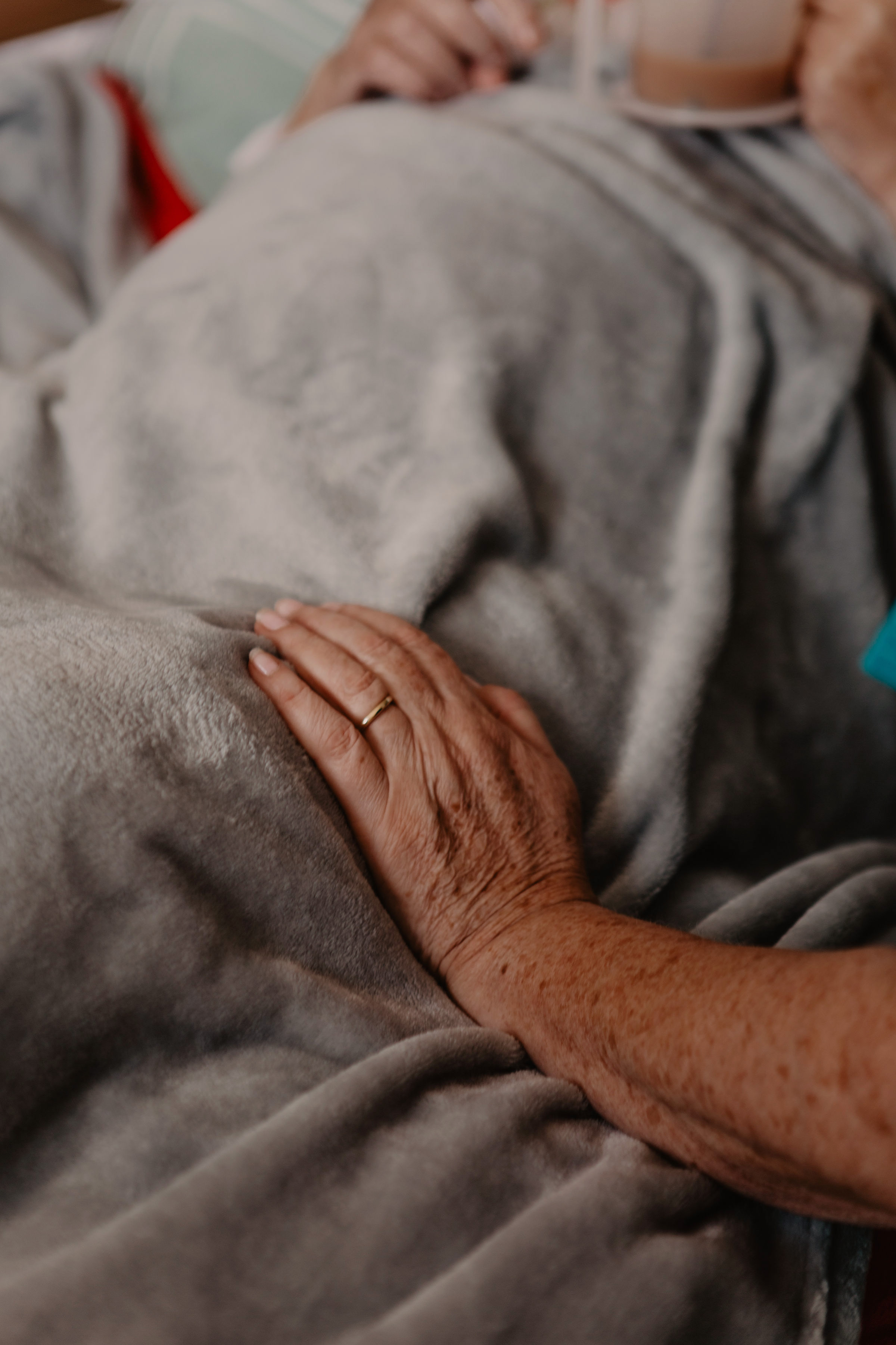 Female healthcare assistant, with her hand resting on a grey blanket of a patient in a hospital bed