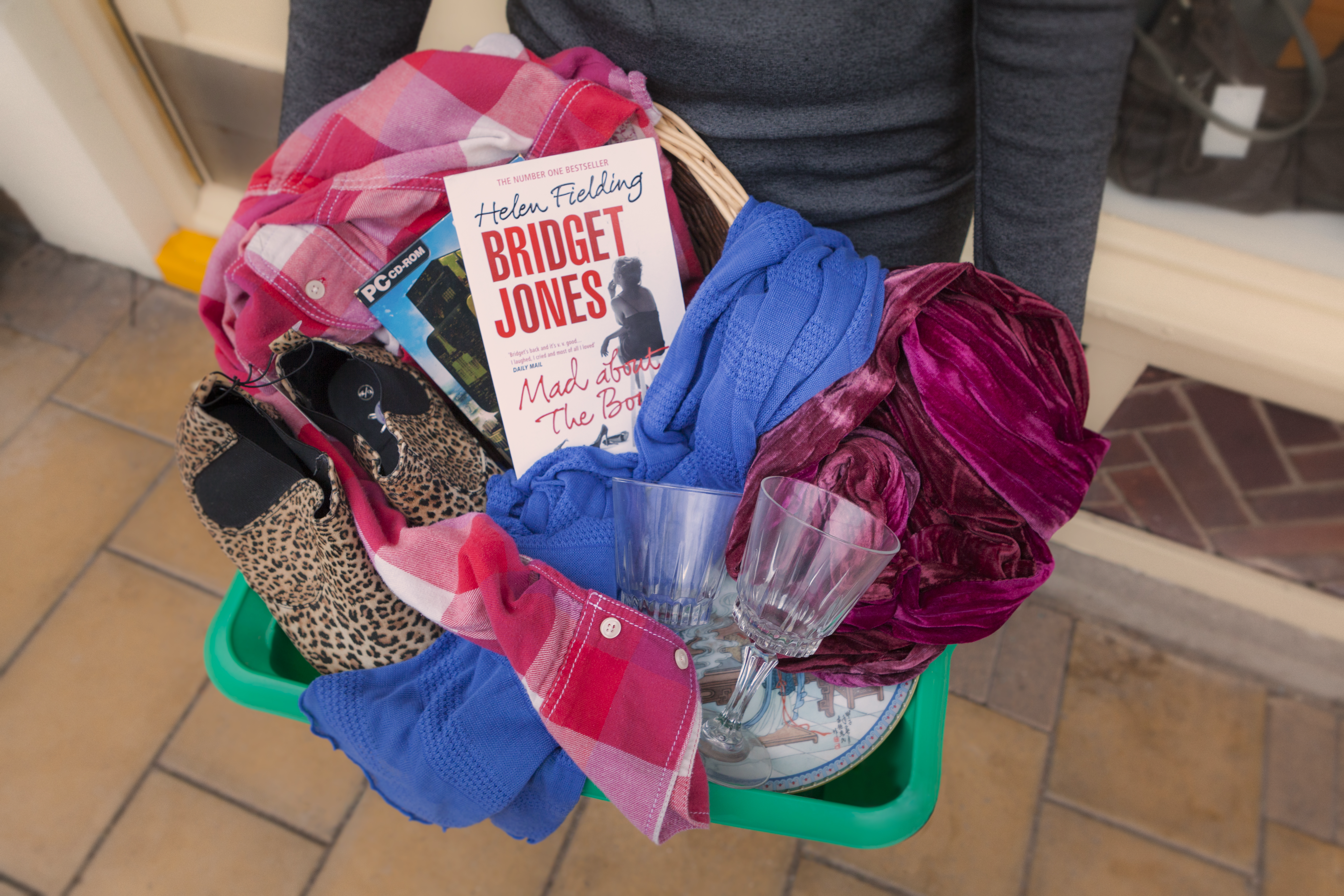 Female in a grey top holding a green box filled with donations - wine glasses, leopard print boots, books and clothing